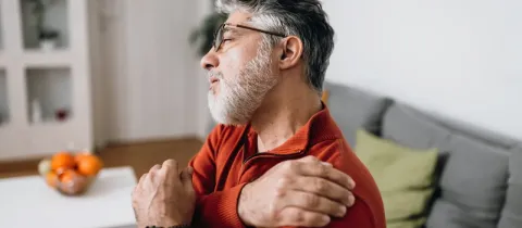 Un homme barbu aux lunettes, le visage grimaçant de douleur, se tient l'épaule.