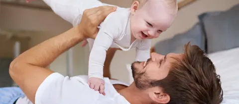 Un père heureux joue avec son bébé, le tenant au-dessus de lui sur le lit.
