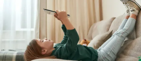 Une petite fille est allongée sur un canapé, les pieds en l'air, regardant une tablette.