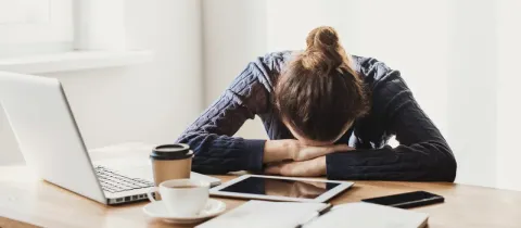 Une femme épuisée, la tête posée sur ses bras, devant son ordinateur portable et des documents.