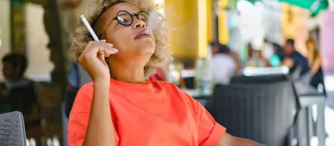 Une femme aux cheveux bouclés et lunettes fume une cigarette, exhalant de la fumée en arrière-plan flou d'un café.