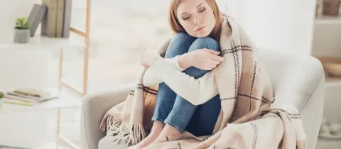 Une jeune femme triste, emmitouflée dans une couverture, assise seule sur un fauteuil.
