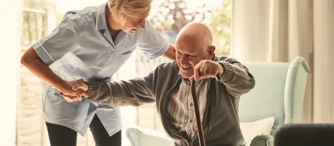 Une infirmière souriante aide un homme âgé à se lever de son fauteuil avec une canne.