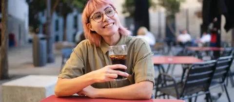 Une jeune femme aux cheveux roses et lunettes souriante, sirotant une boisson sur une terrasse de café.