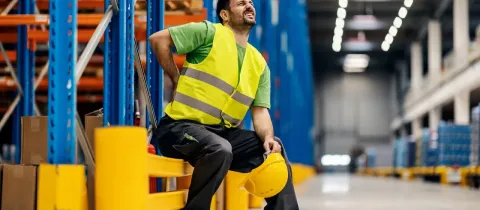 Un travailleur en gilet de sécurité jaune s'assoit dans un entrepôt, se tenant le dos, visiblement en souffrance.