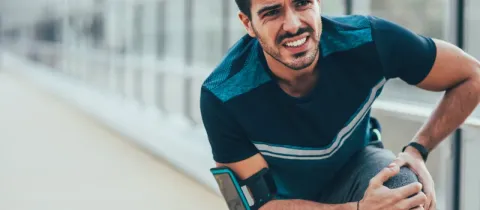 Un homme en tenue de sport grimace et se tient le genou, exprimant une douleur intense pendant l'effort.