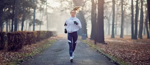 Une jeune femme court sur un chemin bordé d'arbres en automne ou en hiver, vêtue d'une veste blanche.