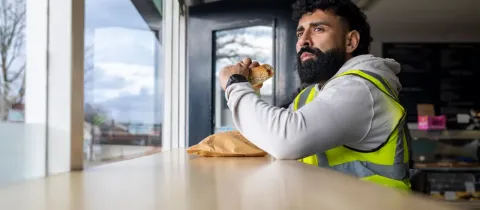 Un homme barbu en gilet de sécurité mange un sandwich près d'une fenêtre, regardant au loin.