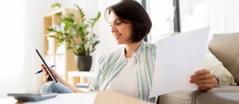 Une femme souriante assise sur le sol chez elle, tenant une tablette et des documents, gérant ses papiers.