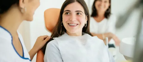 Une adolescente souriante avec un appareil dentaire est assise dans un fauteuil de dentiste, discutant avec le personnel.