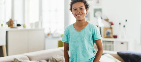 Un jeune garçon aux cheveux bouclés et appareil dentaire sourit et regarde la caméra dans un salon lumineux.