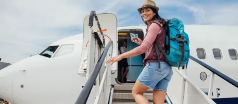 Une jeune femme souriante avec un sac à dos monte l'escalier d'un avion, prête pour le voyage.