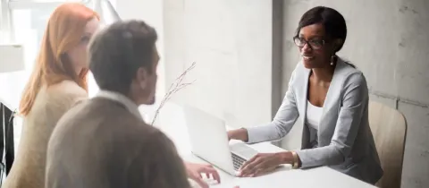 Une conseillère souriante en lunettes discute avec un couple assis à son bureau devant un ordinateur portable.