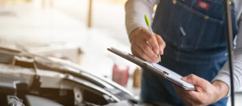 Un mécanicien en salopette écrit sur un presse-papiers, effectuant une inspection sous le capot d'une voiture.