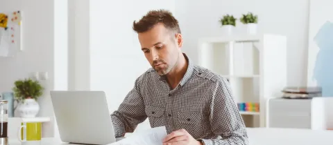 Un homme concentré travaille sur un ordinateur portable, tenant des documents dans un bureau lumineux et épuré.