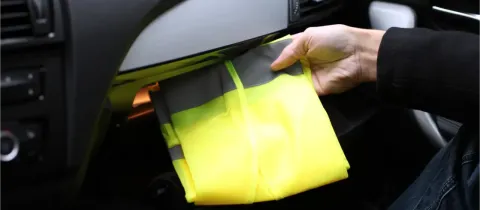 Une main retire un gilet de sécurité jaune fluorescent du vide-poche d'une voiture.