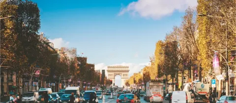 Vue large des Champs-Élysées animés à Paris, avec l'Arc de Triomphe en arrière-plan sous un ciel bleu.