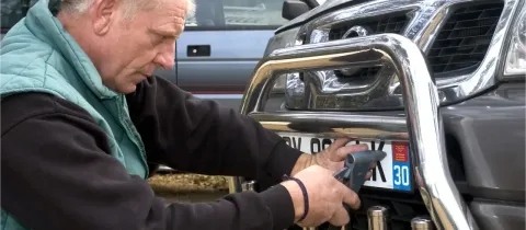 Un homme en veste verte fixe une plaque d'immatriculation sur le pare-buffle avant d'une voiture.