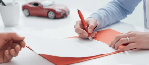 Deux personnes signent des documents d'assurance automobile avec un stylo rouge, une petite voiture rouge est visible en arrière-plan.