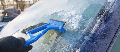 Une main gantée gratte le givre d'un pare-brise de voiture avec un grattoir à neige bleu.