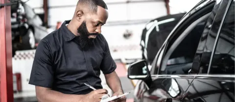 Un mécanicien barbu examine des documents à côté d'une voiture noire dans un garage.