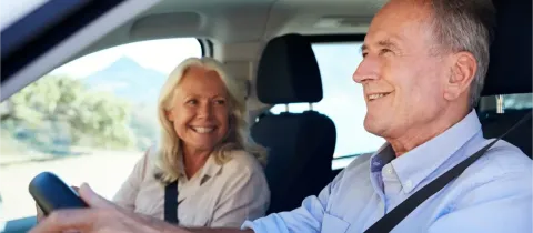 Un couple âgé souriant est assis dans une voiture, l'homme conduit et la femme le regarde.