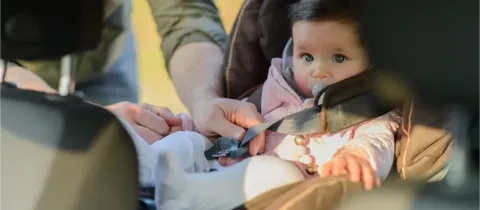 Un adulte ajuste les sangles d'un bébé assis dans un siège auto, le bébé regarde vers l'avant.