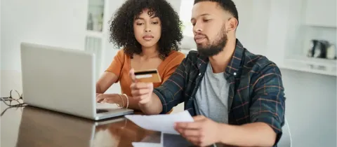 Un couple est assis à une table devant un ordinateur portable, la femme tenant une carte de crédit pendant qu'ils gèrent leurs finances.