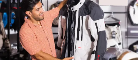 Un homme souriant tient une veste de moto blanche et grise sur un cintre, l'inspectant dans un magasin d'équipement.