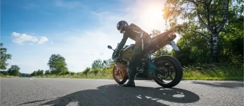 Un motard se penche sur sa moto sportive sur une route de campagne ensoleillée, sous un ciel bleu.