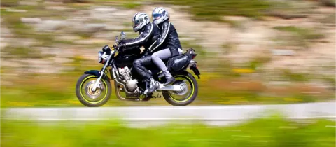 Deux motards en casque et blousons de cuir roulent à vive allure sur une route de campagne verte.