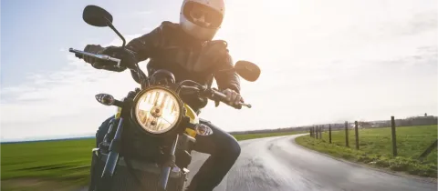 Un motard en blouson de cuir et casque roule sur une route sinueuse en campagne, au soleil couchant.