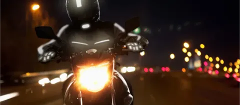 Un motard sur sa moto la nuit, vêtu d'un équipement réfléchissant, avec le phare allumé.