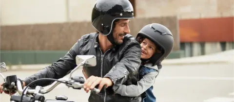 Un père souriant et son enfant en casque noir sur une moto, partageant un moment de complicité.