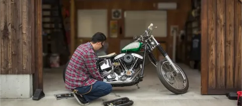 Un homme accroupi répare une moto custom dans un garage en bois ouvert avec des outils.