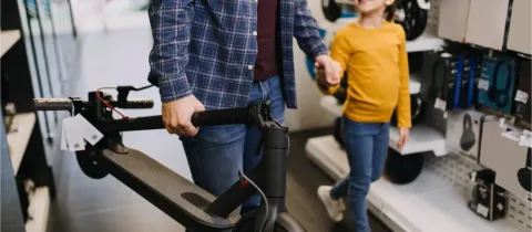 Un homme tient une trottinette électrique tandis qu'une petite fille à lunettes, souriante, lui tient la main dans un magasin.