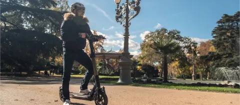Un homme en casque et veste d'hiver se tient à côté d'une trottinette électrique dans un parc ensoleillé.