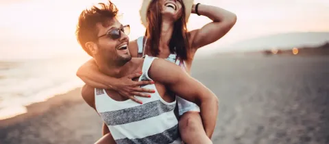 Un couple joyeux s'amuse sur la plage au coucher du soleil, la femme portant un chapeau de paille et riant sur le dos de l'homme.