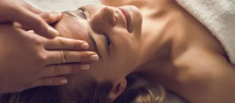 Une personne reçoit un massage relaxant du visage, allongée sous une serviette blanche, les yeux fermés et un sourire apaisé.