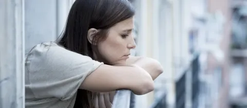 Une femme pensive appuyée sur une rambarde de balcon, regardant au loin, entourée d'immeubles flous en arrière-plan.