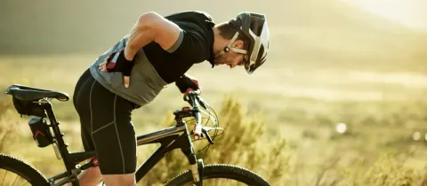Un cycliste en tenue de sport s'appuie sur son vélo, se tenant le dos, dans un paysage ensoleillé et naturel.