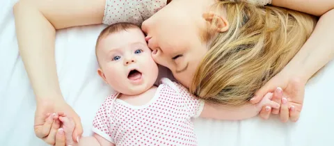 Une femme allongée sur le dos embrasse tendrement un bébé aux yeux bleus sur un lit blanc.