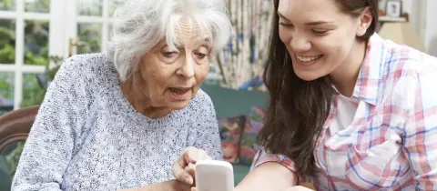 Une jeune femme souriante montre quelque chose sur un smartphone à une femme âgée, assise ensemble dans un salon lumineux.