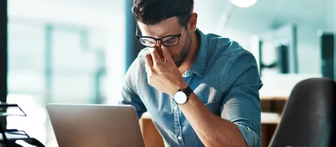 Un homme se frotte les yeux devant son ordinateur, signe de stress/fatigue.