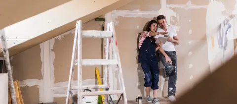 Couple en salopettes de travail planifiant des rénovations dans un grenier, utilisant une tablette près d'une échelle.
