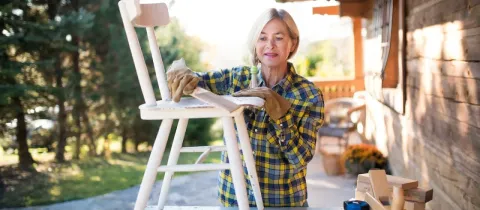Femme mûre ponçant une chaise en bois sur une terrasse extérieure. Elle porte des gants de travail et une chemise à carreaux.