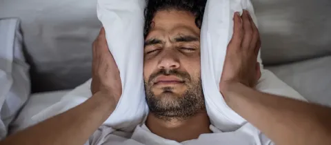 Gros plan sur un homme au lit qui couvre ses oreilles avec des oreillers blancs, l'air irrité par le bruit.