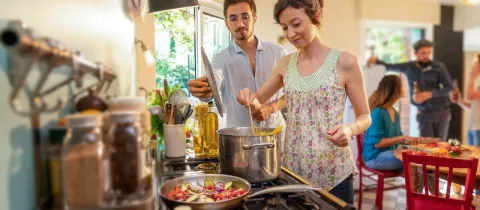 Couple préparant des pâtes et des légumes dans une cuisine ensoleillée, avec des amis en arrière-plan.