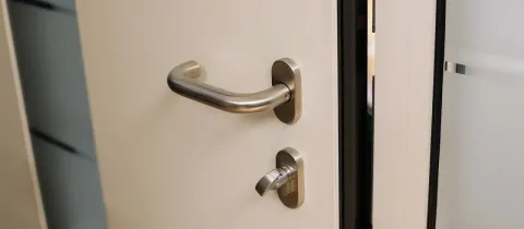 Gros plan sur une porte blanche avec une poignée et une serrure métalliques modernes, un montant vitré est visible à droite.