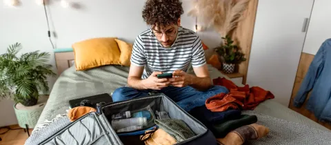 Jeune homme bouclé, valise ouverte devant lui, vérifiant les informations de voyage ou la météo sur son téléphone.